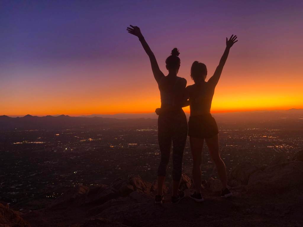 Silhouette of two girls looking over the edge of Camelback Mountain during a vibrant orange and purple sunrise in Phoenix, Arizona