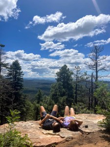 A boy and girl sitting on the edge of a cliff in Arizona, in love and talking about life. Blue skies, white puffy clouds, green pine trees.