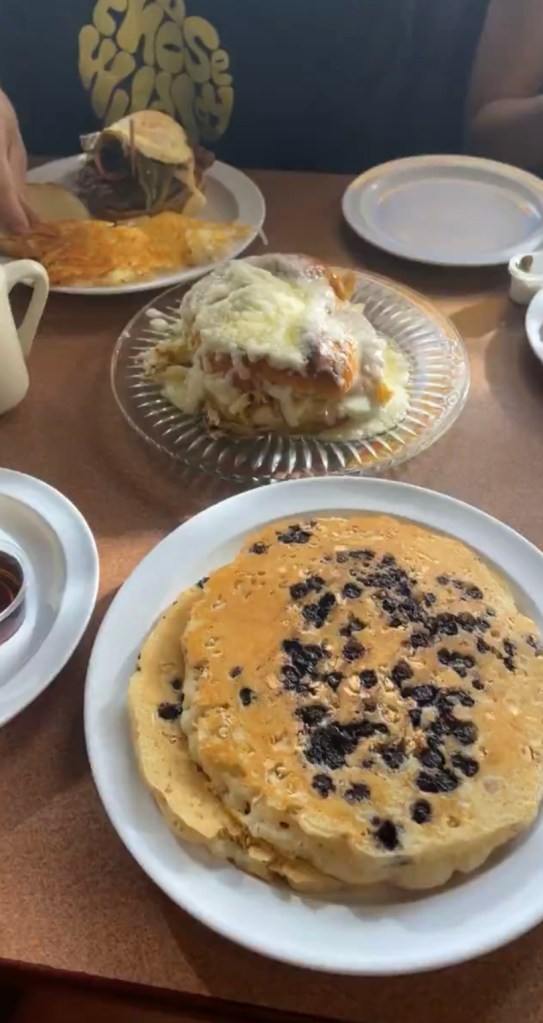 stack of pancakes with granola and blueberries, giant cinnamon roll in the middle of the table, wildjoy sweatshirt in the background