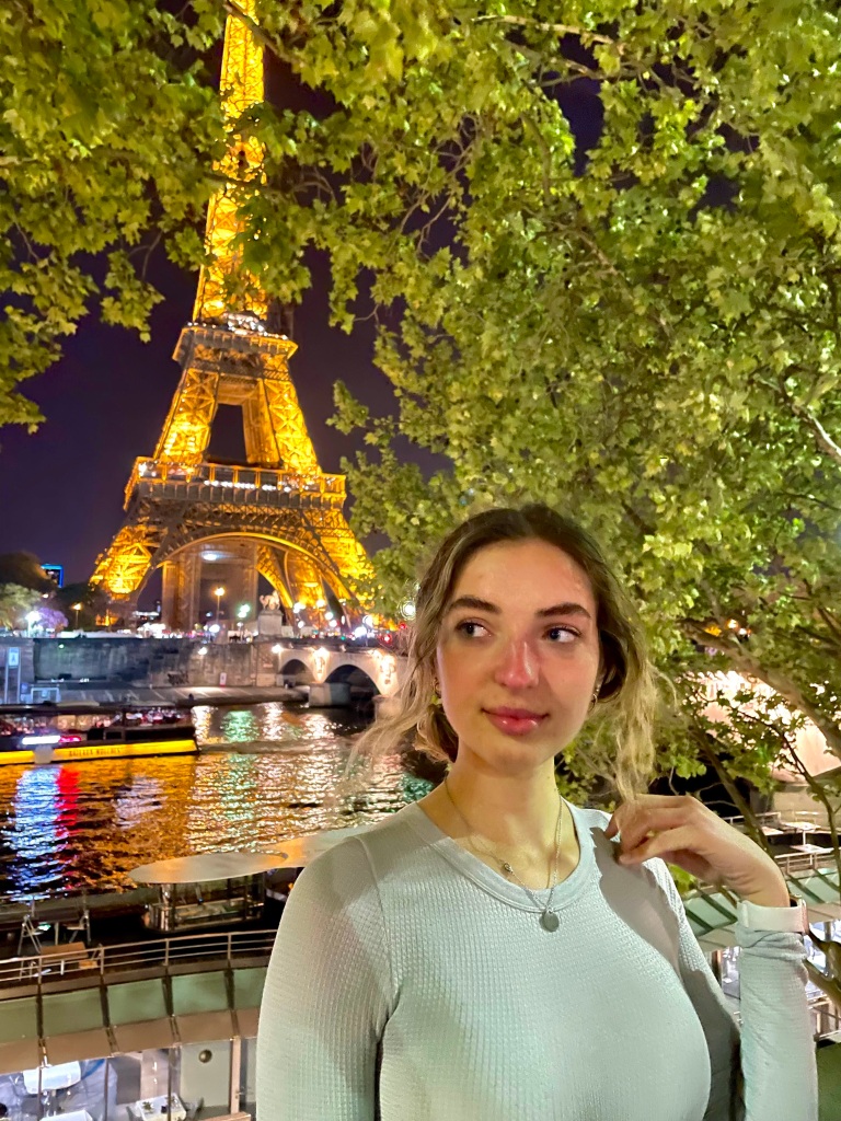 A girl looking off into the distance as she poses in front of the Seine and the Eiffel Tower at night.