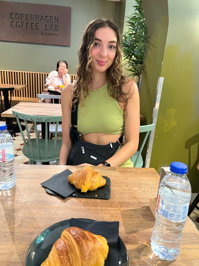girl sitting in a coffee shop in Lisbon, smiling with a croissant, lady in the back photo bombing and smiling