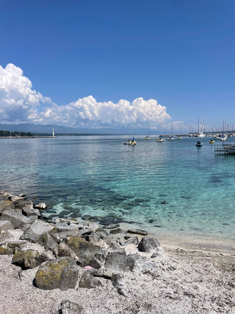 A small sandy beach on the edge of Lake Geneva with super clear, light blue waters and mountains in the distance in Geneva, Switzerland