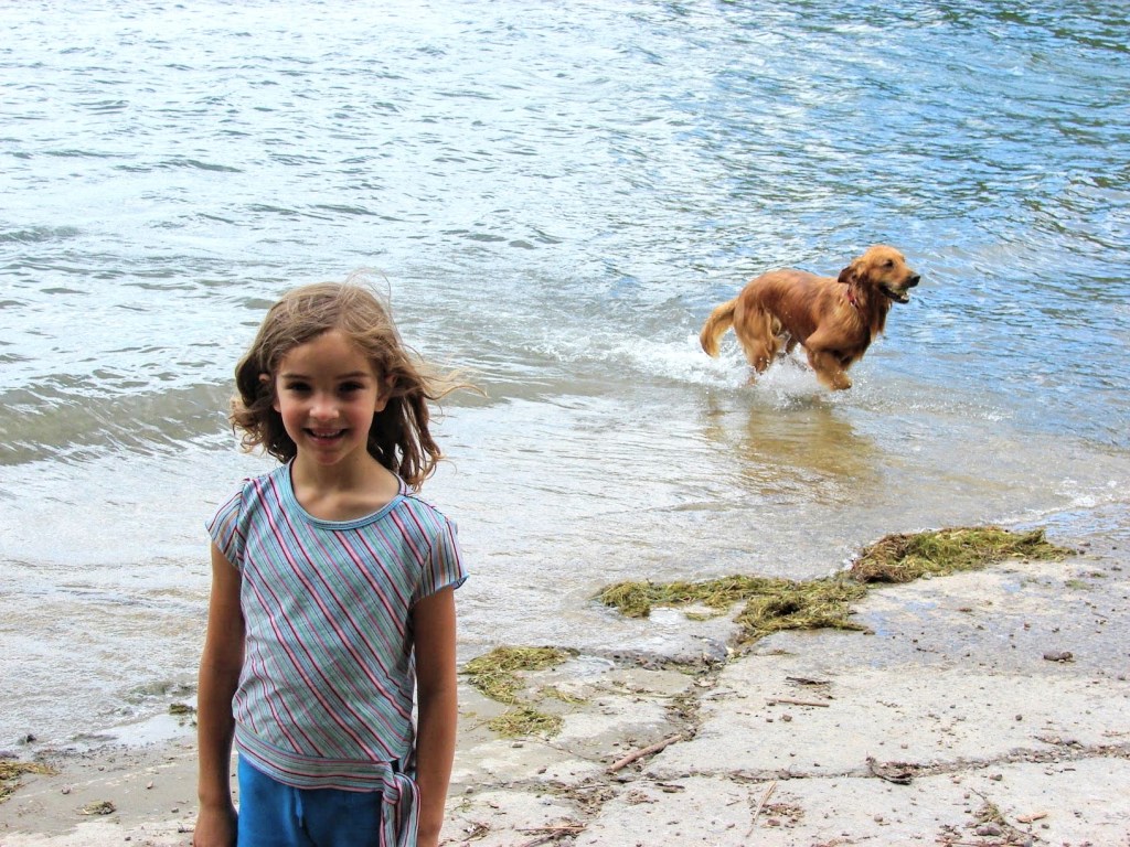 A young girl standing and smiling for the camera in front of a lake, and behind her a golden retriever puppy is running through the water with a tennis ball in his mouth
