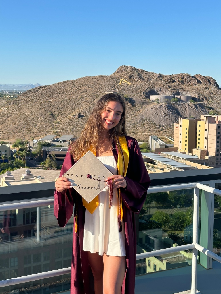 A recent college graduate of Arizona State University smiling while holding her graduation cap that is decorated and says "Next stop, France" in front of A Mountain in Tempe, Arizona