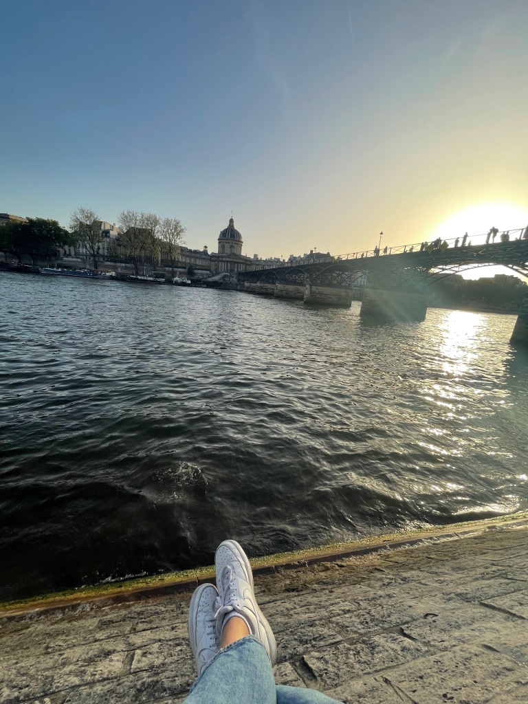 feet swaying off of the river bank while a girl watches the sunset on the Seine in Paris, France next to Pont Neuf