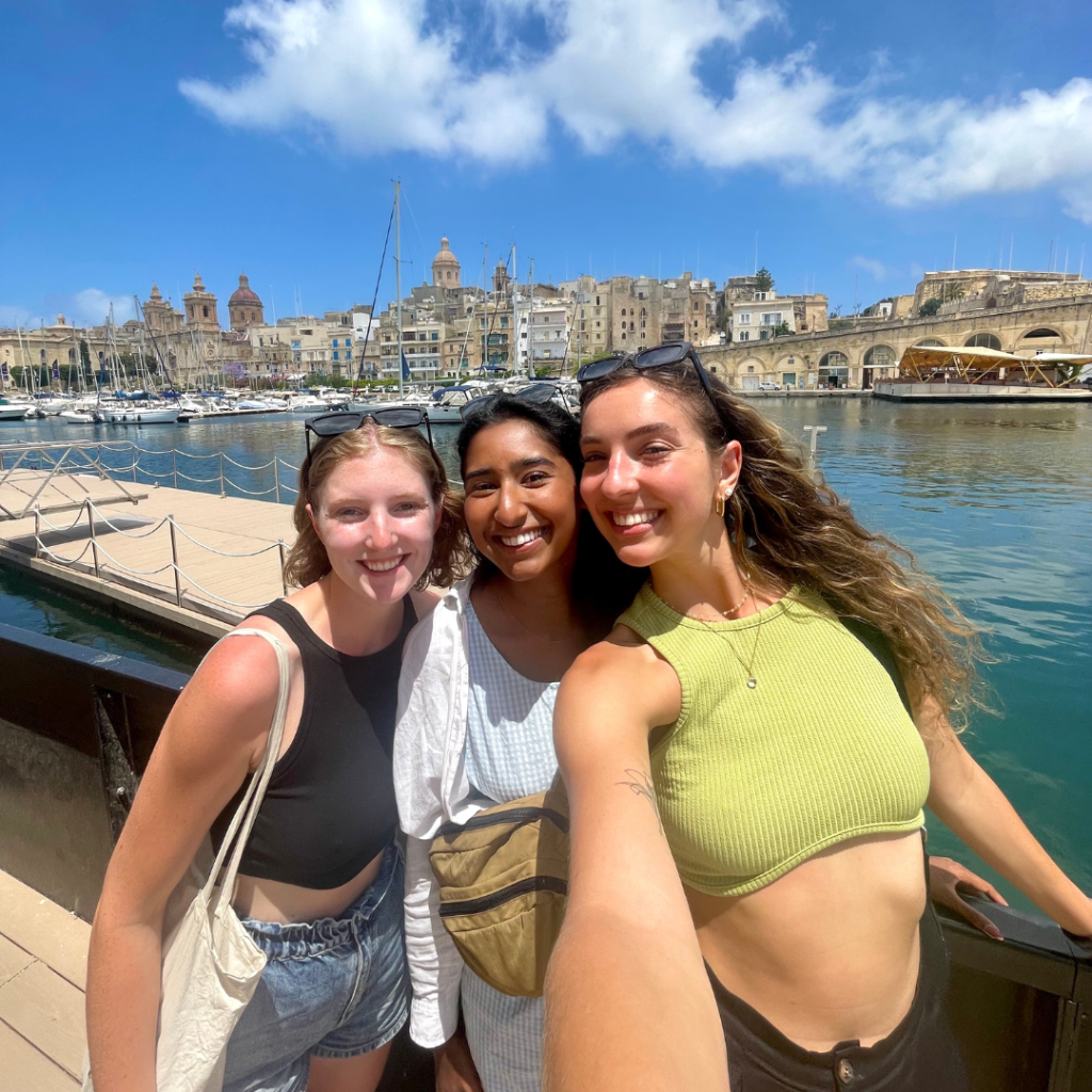 Three best friends smiling for a .5 selfie on the island of Malta on a sunny day with the port in the background and beautiful limestone buildings