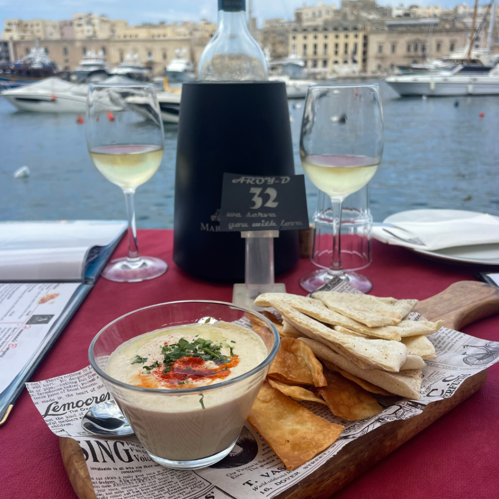 A plate of freshly made pita chips with homemade hummus, a bottle of wine and two glasses, on an outdoor patio with a view of the marina at a Mediterranean restaurant in Malta