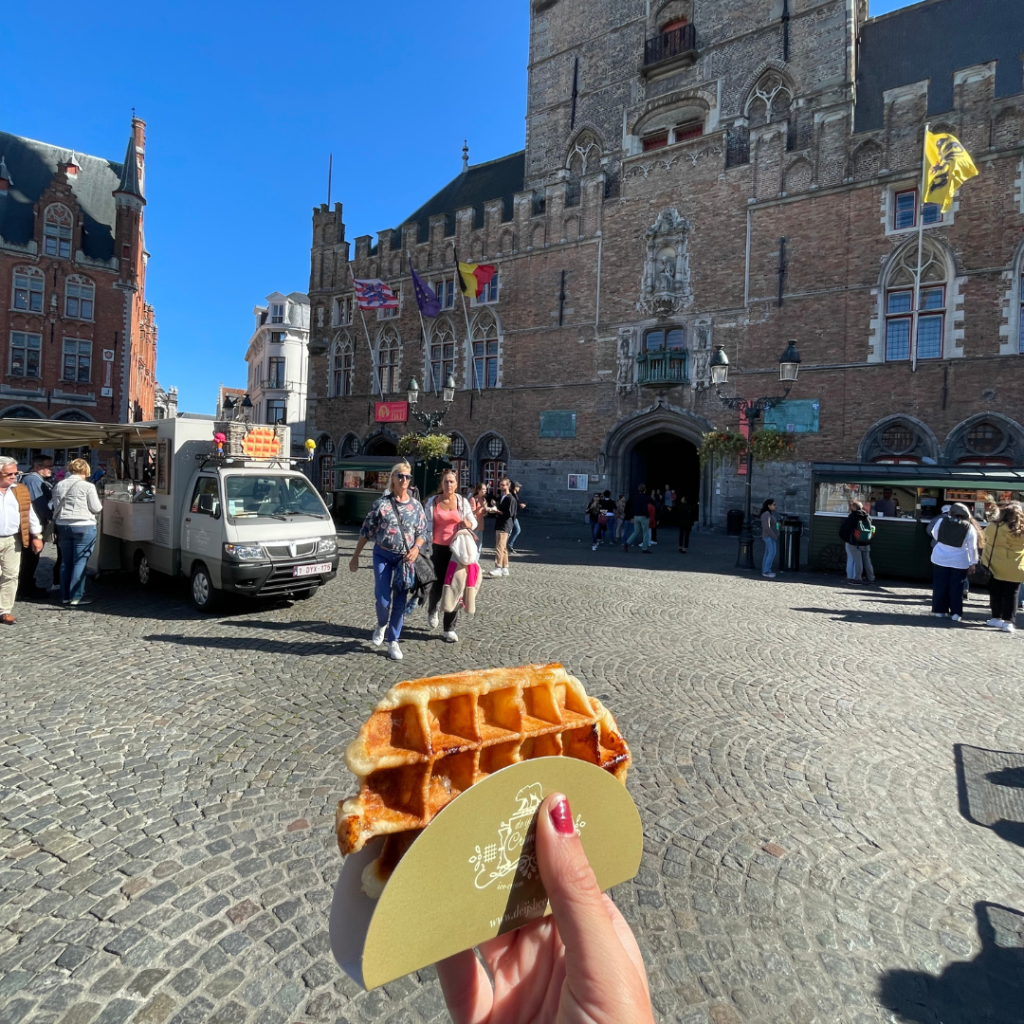 A hand holding a liege waffle in a paper cone in the brick streets of Bruge, Belgium on a sunny day