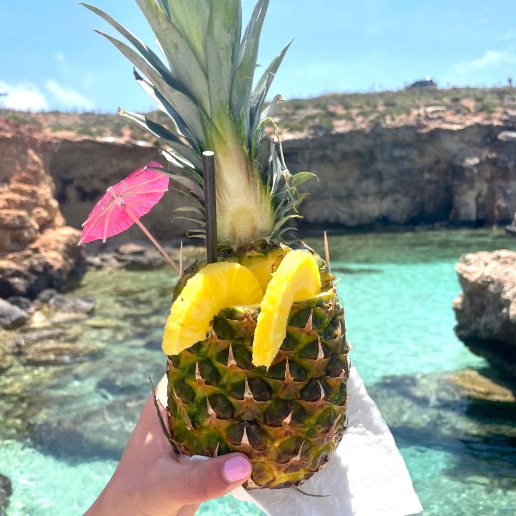 A hand holding a piñacolada in a pineapple in front of the crystal clear lagoon/sea off of the island of Malta