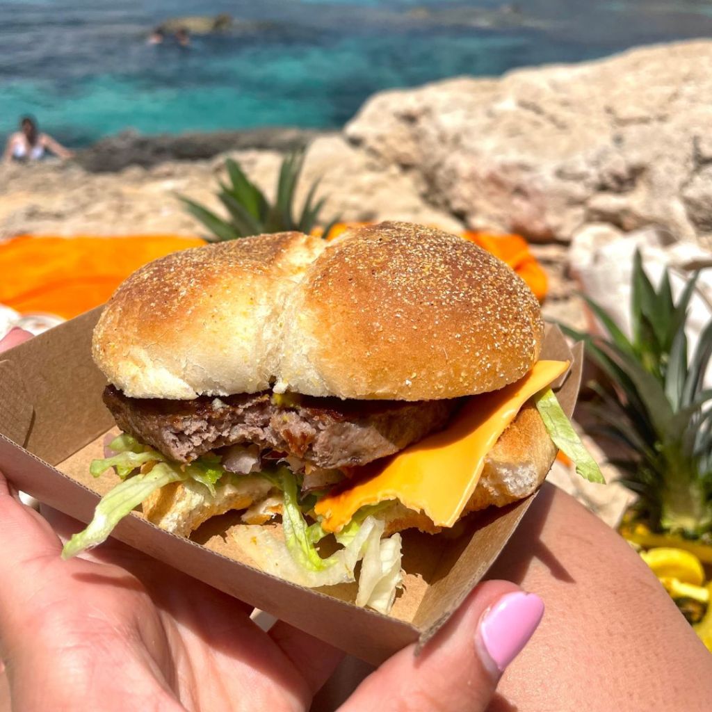 A hand holding a cheese burger in front of the crystal clear lagoon/sea off of the island of Malta