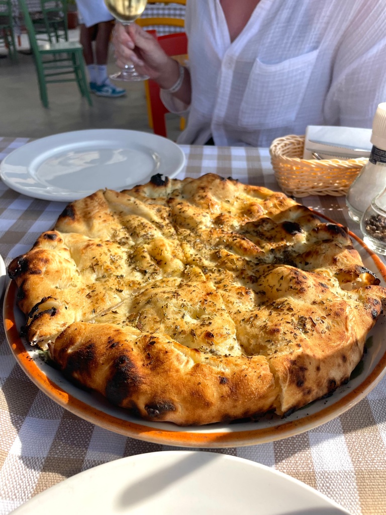 In the foreground, a perfectly cooked, golden focaccia seasoned with olive oil and herbs, reflecting perfectly off of the sun during golden hour during an apero on the island of Malta. A woman in the background elegantly holding a glass of white wine
