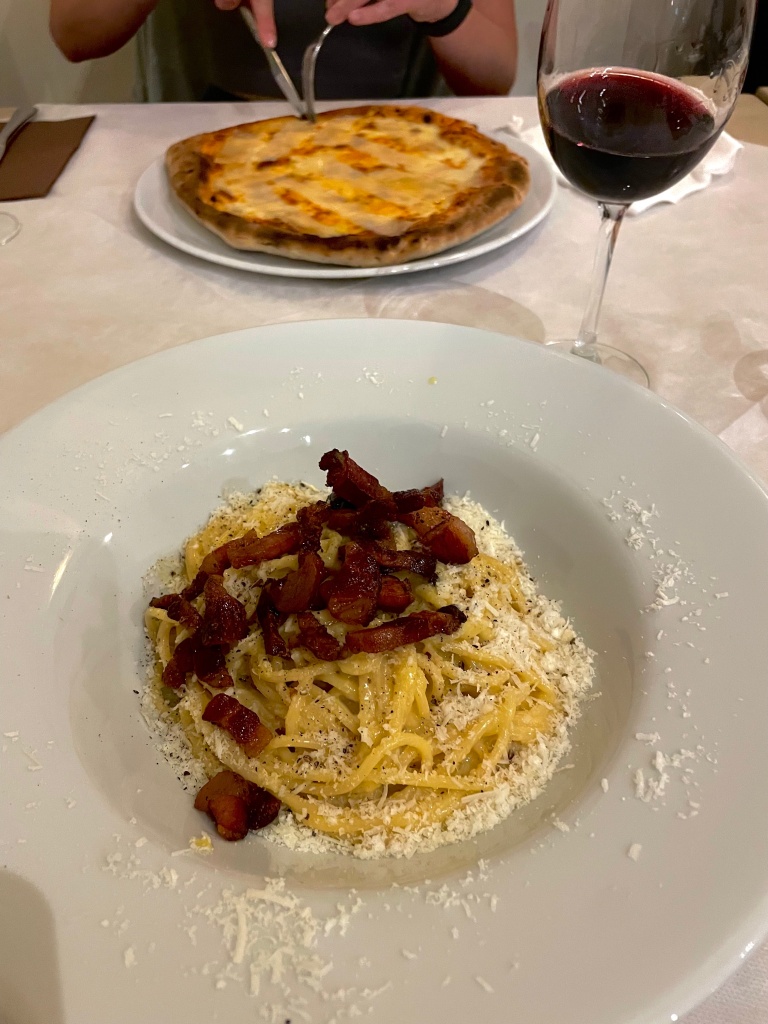 Pasta Carbonara topped with bacon and parmesean cheese in a large, white bowl, with someone slicing their pizza in the background, accompanied by a glass of red wine at an Italian restaurant in Malta.
