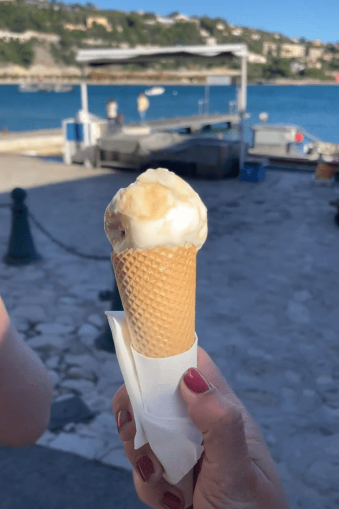 A hand holding salted caramel gelato in a cone in front of the Mediterranean Sea near Nice, France