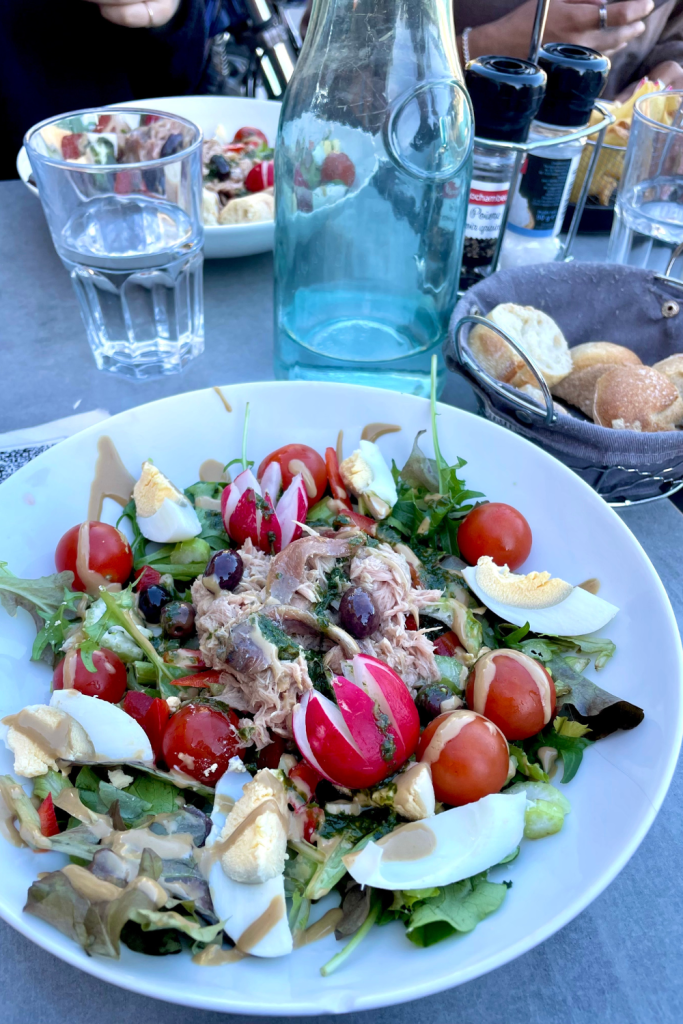 Niçoise salad topped with tuna, hard boiled eggs, radish, tomatoes, and potatoes, with a basket of bread and other dishes in the background, at a restaurant in Villefranche sur Mer, France