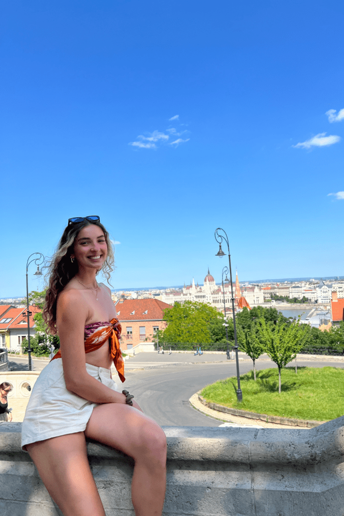 A tall woman smiling for a photo in Budapest with the Parliament in the background and vibrant green trees in the foreground on a sunny summer day.