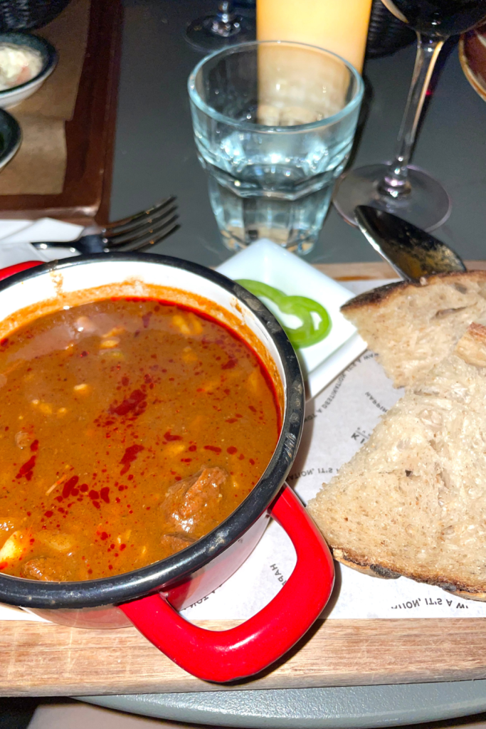 Traditional goulash in a cozy red bowl served with a side of freshly made bread, with a glass of wine in the background at a restaurant in Budapest, Hungary.