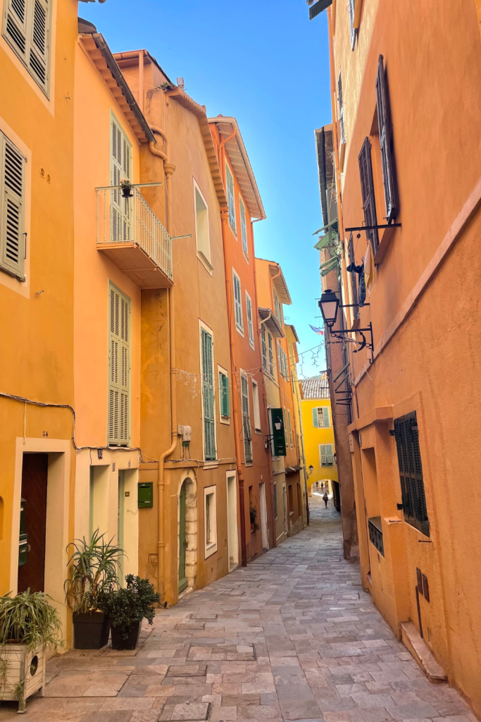 Exploring the narrow streets of Villefranche sur Mer near Nice, France. With vibrant orange and yellow buildings and a bright blue sky peaking out at the top. 