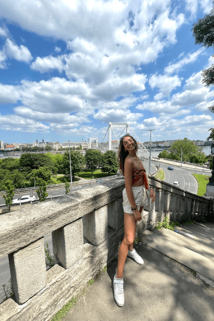 A tall girl smiling and posing in front of the Erzsébet Bridge and Danube River in Budapest, Hungary, on a sunny day in the summer.