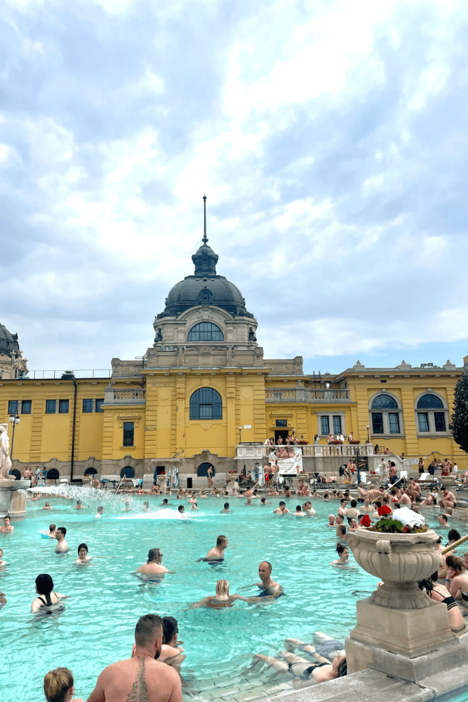 The Szechenyi Thermal Baths featuring people relaxing in the spa with clear, light blue water a cloudy day in Budapest, Hungary.
