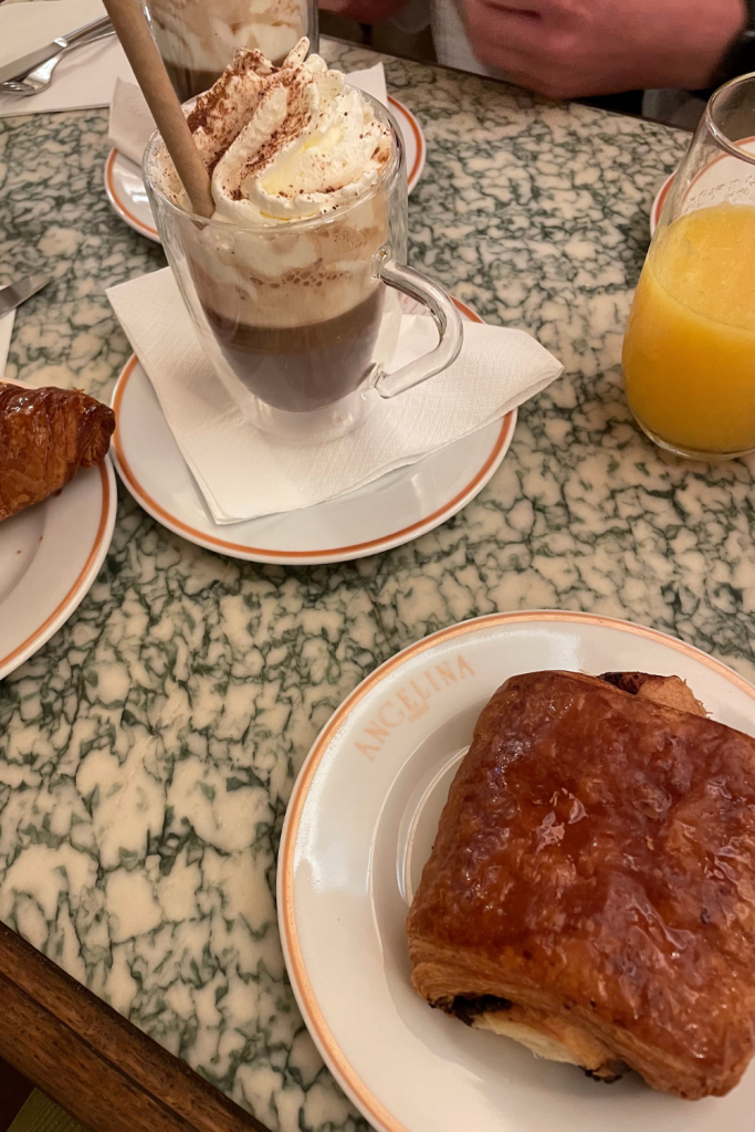 A perfectly flakey and buttery pain au chocolate with a mocha hot chocolate topped with whipped cream in the background at Angelina's in Paris