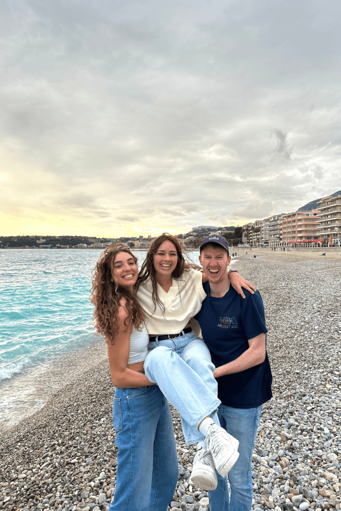 Two friends holding up a third fried for a picture at the beach while traveling to the south of France