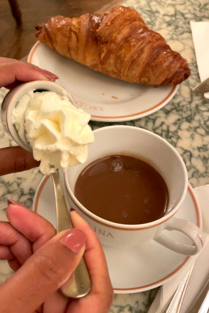 A woman's hand topping the famous hot chocolate with whipped cream at Angelina's in Paris with a buttery flakey croissant in the background