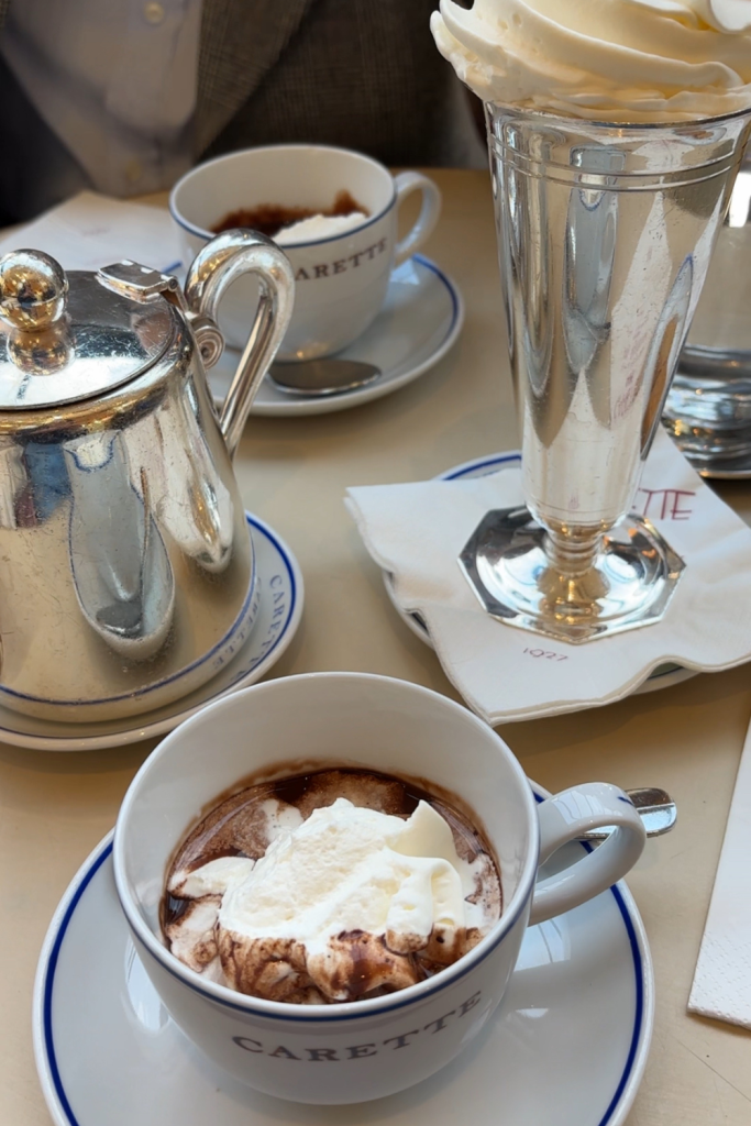 Two mugs filled with velvety rich hot chocolate, topped with whipped cream, and a tea pot filled with more hot chocolate at Carette in Paris