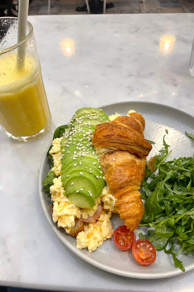 Brunch at a café in Nice, France, featuring a delicious croissant filled with scrambled eggs, avocado, and sliced turkey. Garnished with a side salad. A freshly made juice in the background.