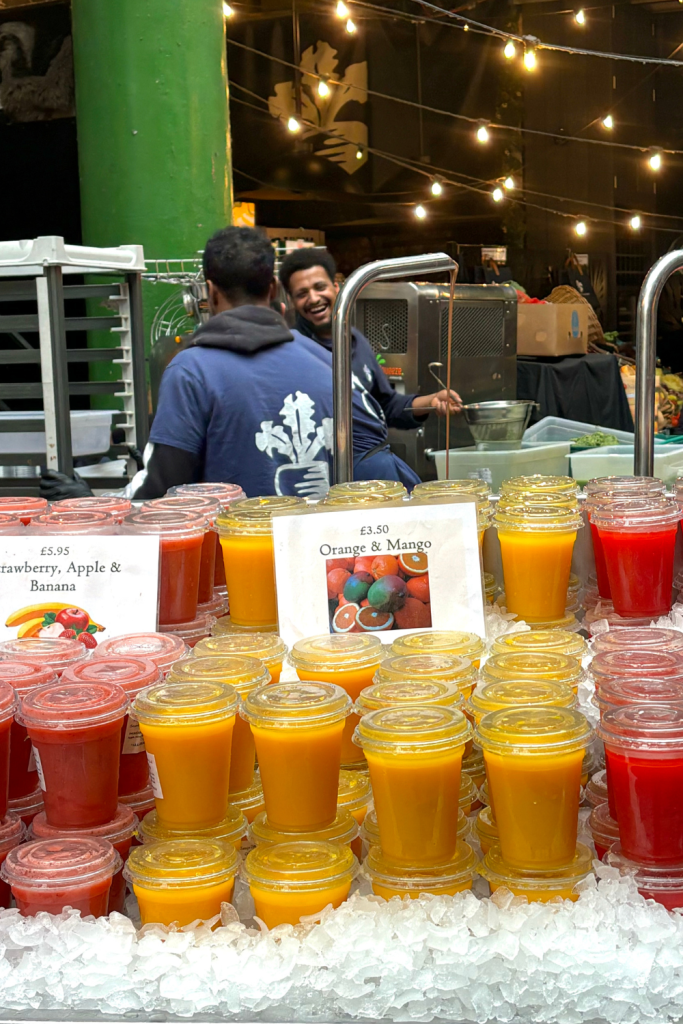 A produce stand at Borough Market in London displaying a wide variety of freshly squeezed juices, featuring a mango orange juice and strawberry, apple, and banana juice. With two men in the background laughing as they cover strawberries with chocolate.