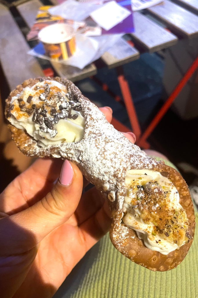 A hand holding a homemade large cannoli topped with chocolate chips and pistachios from a gelateria in Florence, Italy