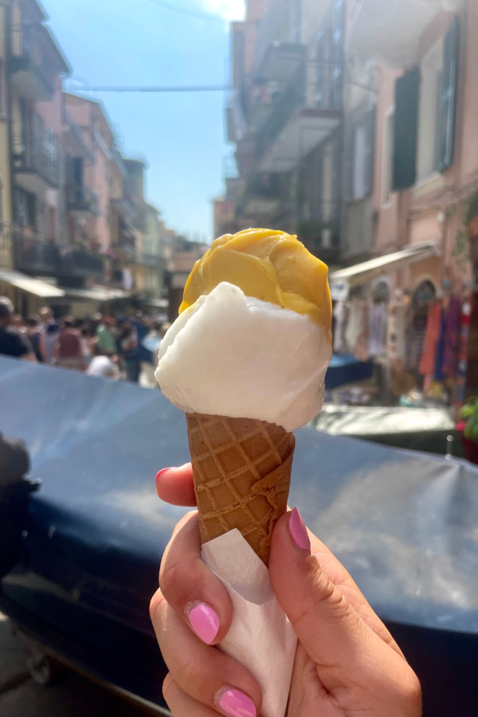 A woman's hand holding a cone filled with the most refreshing and tasty mango and coconut gelato found in Cinque Terre, Italy on a sunny day in the summer