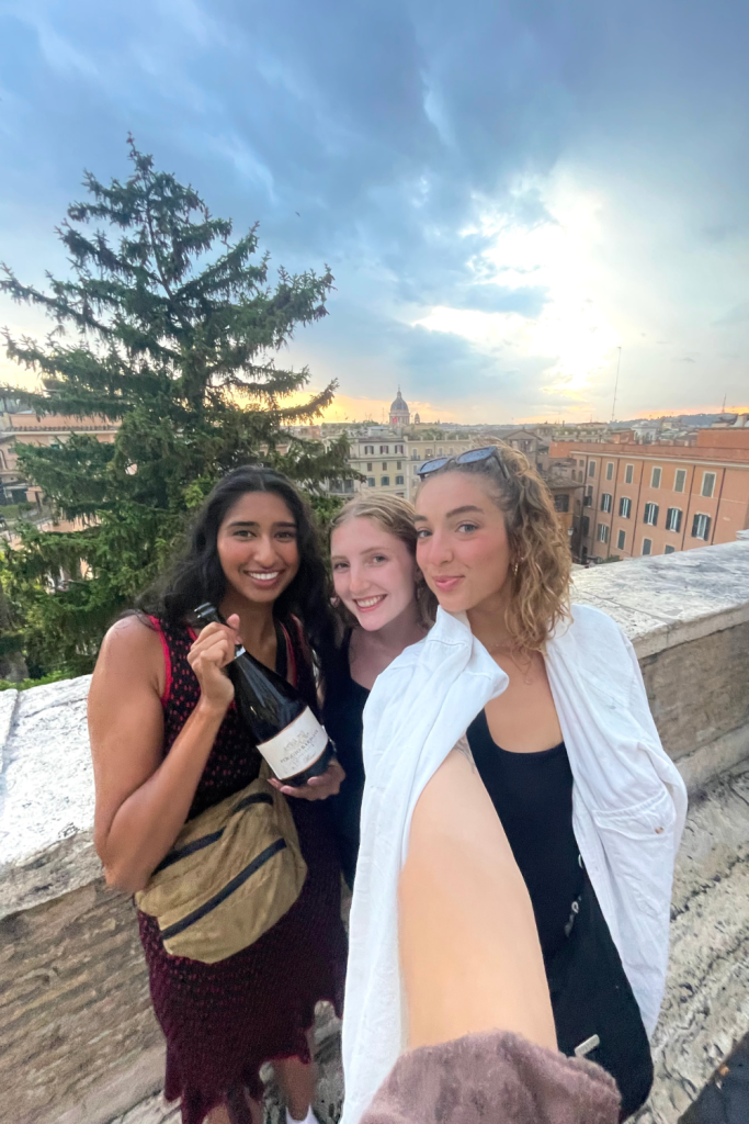 Three best friends enjoying the beautiful and light yellow and blue sunset at the top of the Spanish Steps in Rome, Italy, holding a bottle of Italian Rosé from Tuscany.