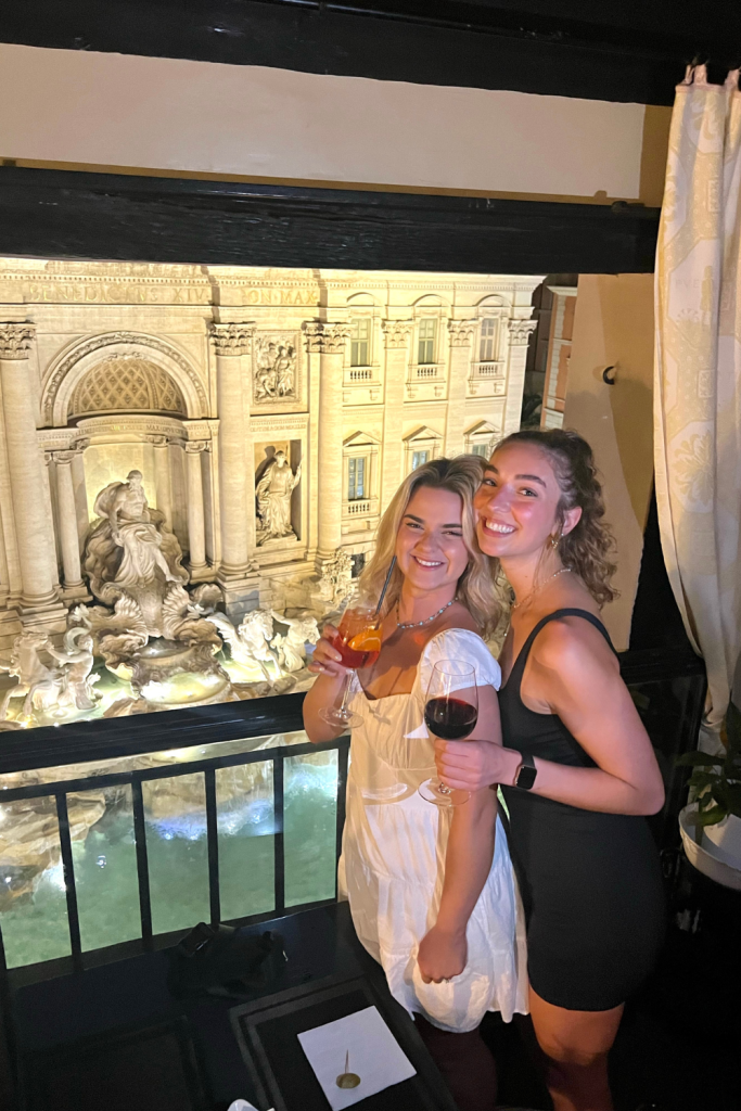 Two best friends dressed up for the night out, holding an Aperol Spritz and glass of red wine, at an exclusive restaurant above the Trevi Fountain in Rome, Italy