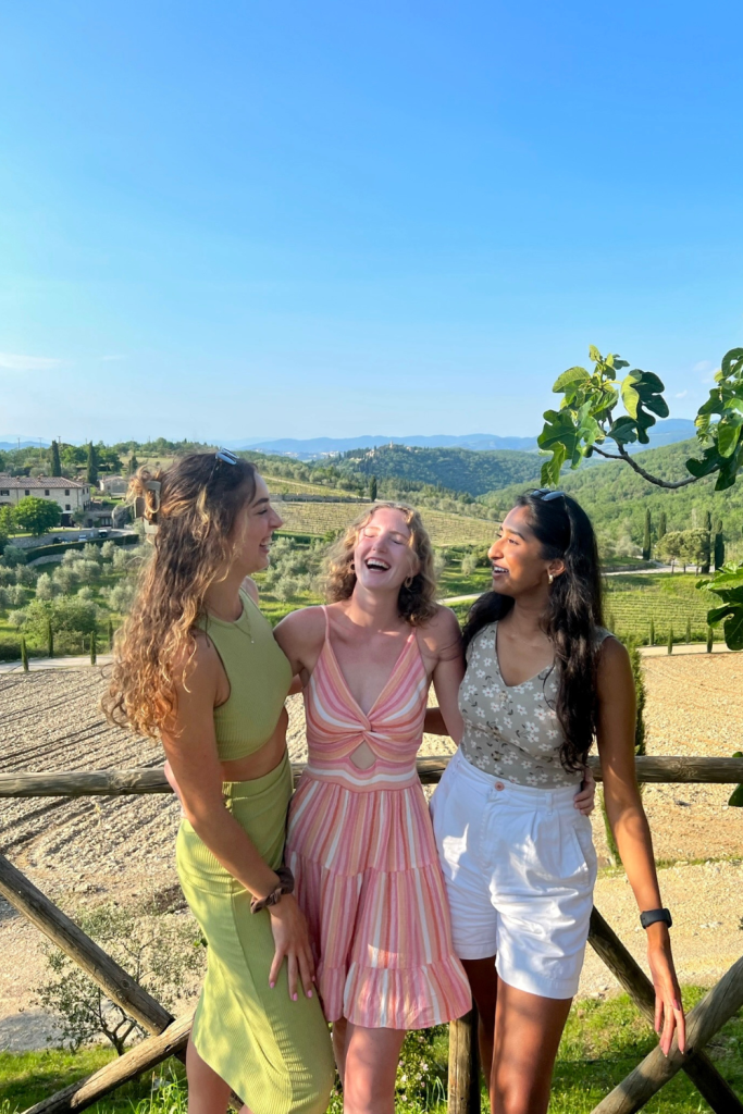 Three best friends laughing together in front of rolling green hills while on a wine tasting tour in Tuscany, Italy