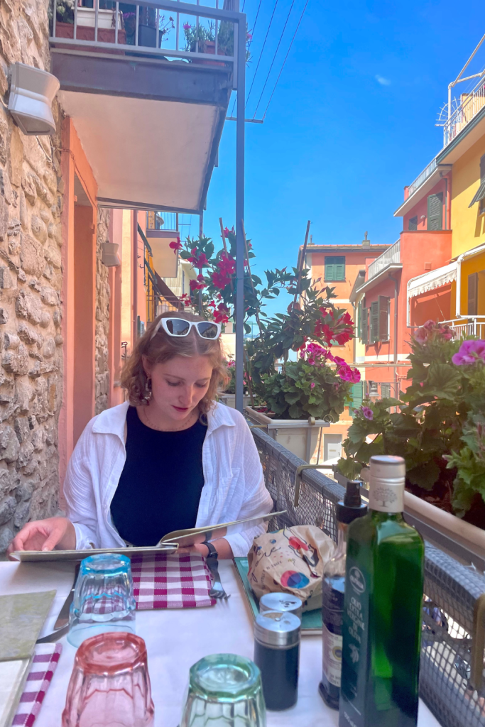 A girl looking at the menu on the balcony at an Italian restaurant in Cinque Terre, with a bright blue sky and colorful buildings in the background