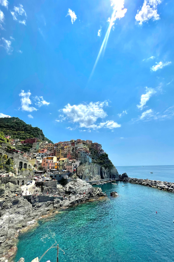 A gorgeous view of the colorful buildings on the cliffside of Manarola in Cinque Terre, Italy on the sunniest day with bright blue turquoise waters to the right.