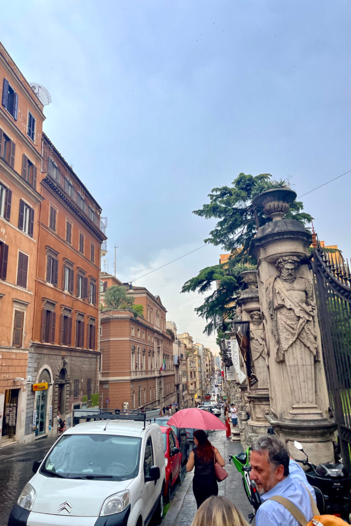 Walking down the streets of Rome, Italy towards the Spanish Steps after a rainfall. Light blue clouds with the sun peaking through, and a lady walking with a red umbrella.