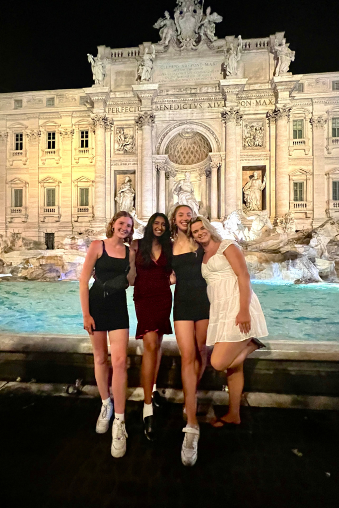 Four gorgeous friends dressed up for a night out, laughing and smiling in front of the Trevi Fountain in Rome, Italy at night, with the water lit up crystal blue