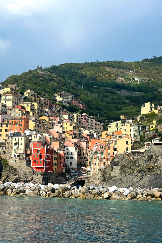 A view from the sea of the beautiful colorful buildings of Riomaggiore in Cinque Terre, Italy, with the rolling green hills in the background on a clear, blue sky day.