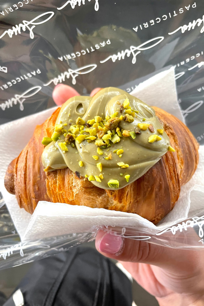 Hand holding a pistachio cream filled croissant, topped with the decadent pistachio cream and pistachio crumbles. From a bakery in Rome, Italy
