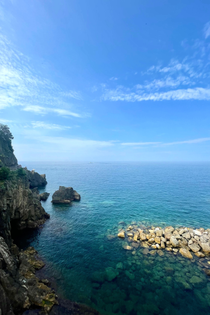 Gorgeous view of the Ligurian Sea from Riomaggiore in Cinque Terre, Italy, with deep turquoise waters, rocks, and a cliff slide on a clear blue sky day