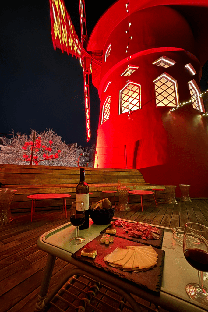 A planche of charcuterie, fromage, and a bottle of wine with the windmill of Moulin Rouge in the background from a rooftop bar in Paris, France.