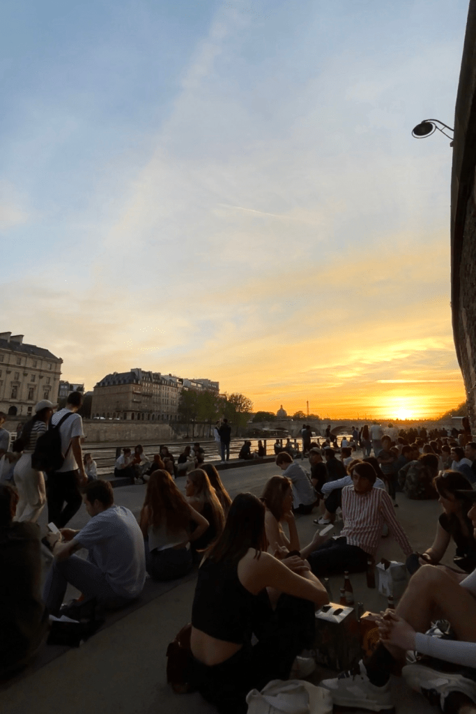 Many groups of friends sitting along the quai of the Seine in Paris, France during a beautiful spring night and a gorgeous, orange and bright yellow sunset.