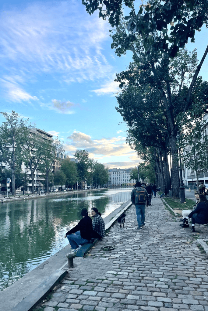 Walking along the quiet Canal Saint-Martin in the 10th arrondissement in Paris on a sunny, fall day. A cute couple is sitting along the canal, enjoying the views and a man is ahead walking his small dog.