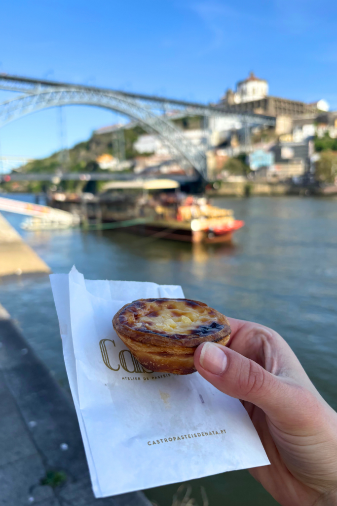 A woman's hand holding a perfectly baked pastel de nata in front of the Douro River in Porto, Portugal.