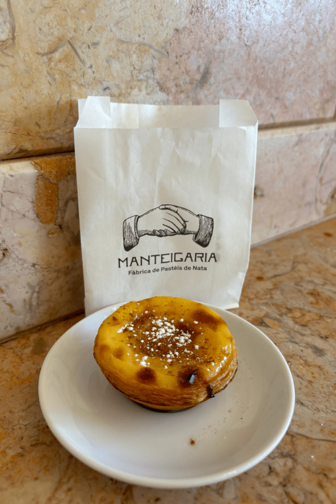 A perfectly golden pastel de nata topped with powdered sugar and cinnamon on a mini white plate from a Pastelaria in Porto, Portugal.