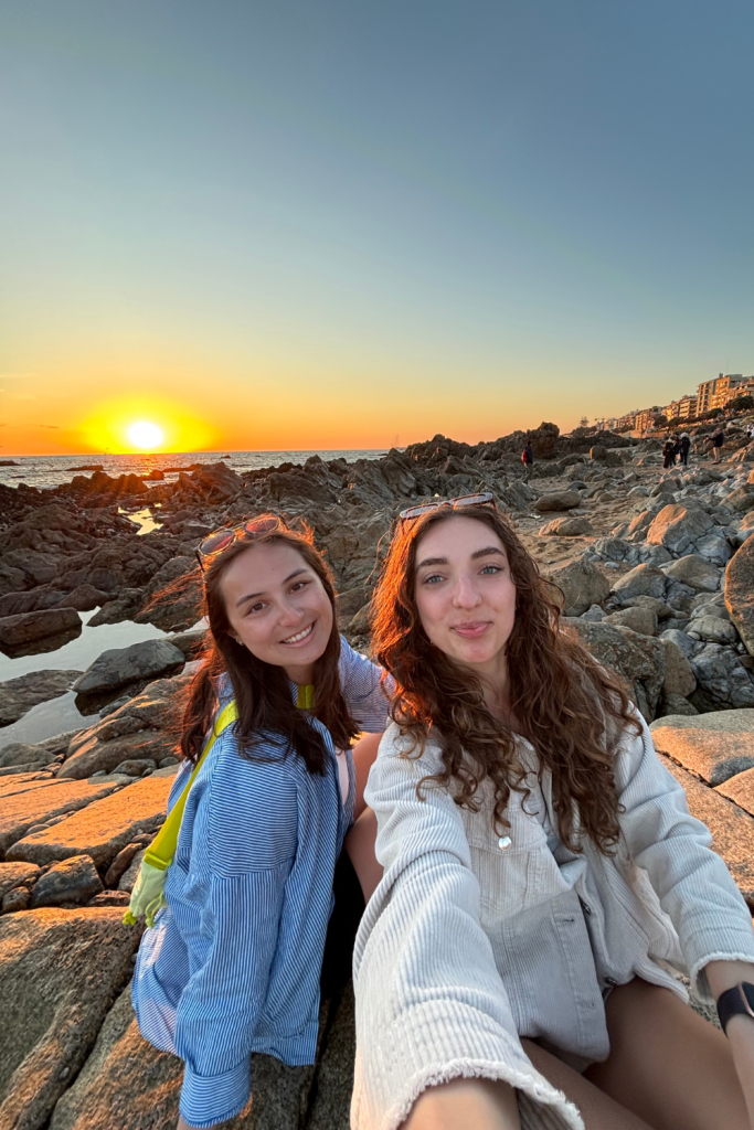 Two solo travelers who became friends while traveling smiling for a photo with the sunset in the background at the beach near Porto, Portugal.
