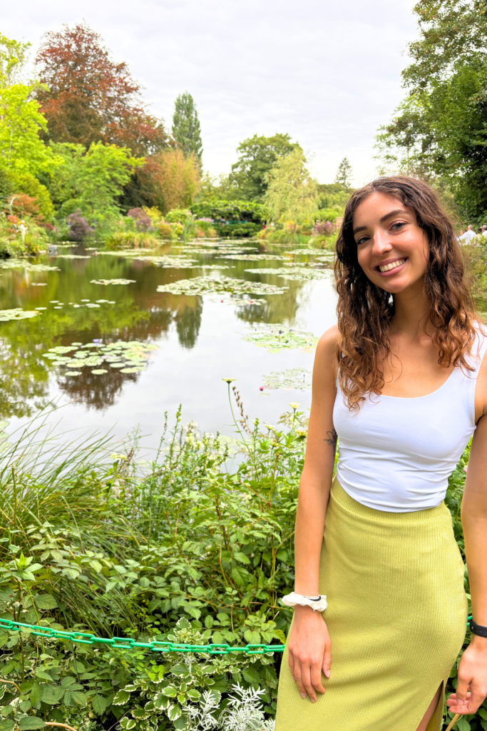 A young traveler smiling in for a photo in front of the famous pond and garden at Claude Monet's house in Giverny, France, surrounded by a ton of greenery and lilypads.