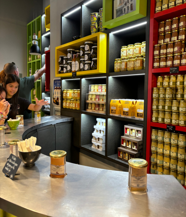 A wall of Dijon mustard on display in a family run mustard shop in France with a sampling counter in front and a girl reaching for another sample.