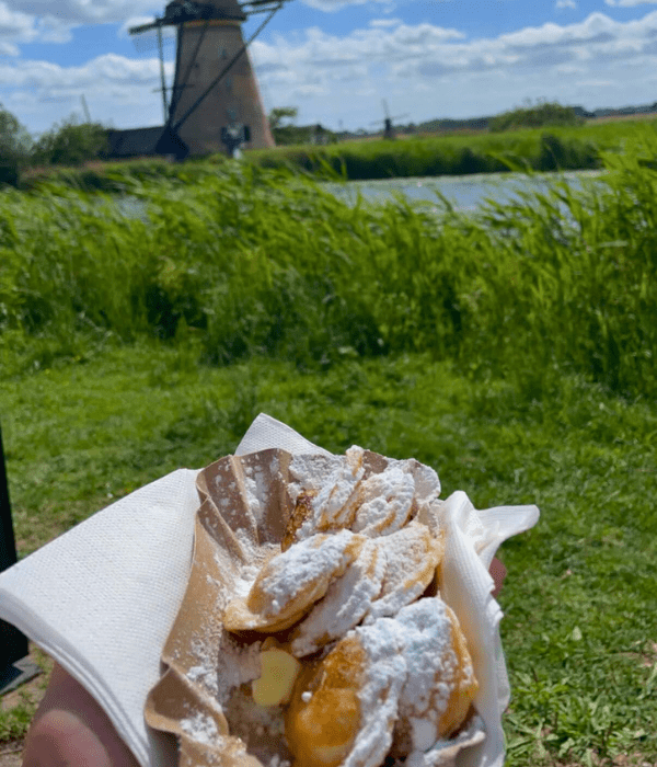 A plate of freshly made mini Dutch pancakes, or Poffertjes topped with powdered sugar, in front of the windmills in Kinderdijk